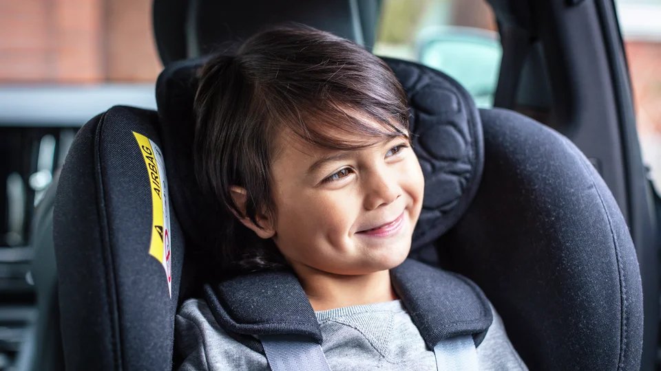 Pojke i bakåtvänd bilbarnstol tittar leende ut genom bilrutan.