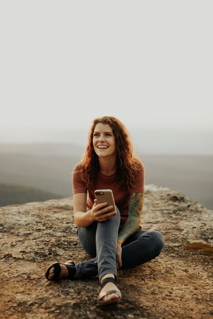 En ung kvinne iført rustrød t-skjorte, grå olabukse og sandaler sitter ute i naturen på toppen av et berg og smiler til kameraet med mobilen i hånden. Hun har krøllete rødlig hår, et pent smil og har en tatovering på sin venstre arm.