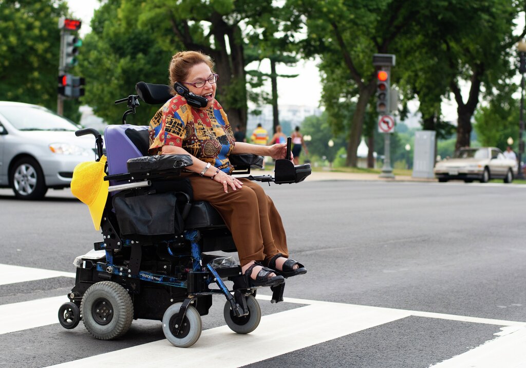 Judy Heuman in motion, crossing a pedestrian crossing. She sits in an electric wheelchair and wears brown trousers, sandals and a blouse in many different variations of primary colours and patterns. Between her shoulder and her cheek she has the mobile phone she is talking on.