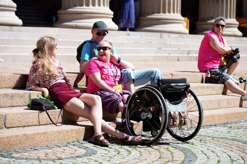 Illustrasjonsfoto av en funksjonshemmet kvinne som sitter i solen i en trapp omgitt av assistenten sin på den ene siden, og rullestolen på den andre. Rundt dem sitter andre mennesker.