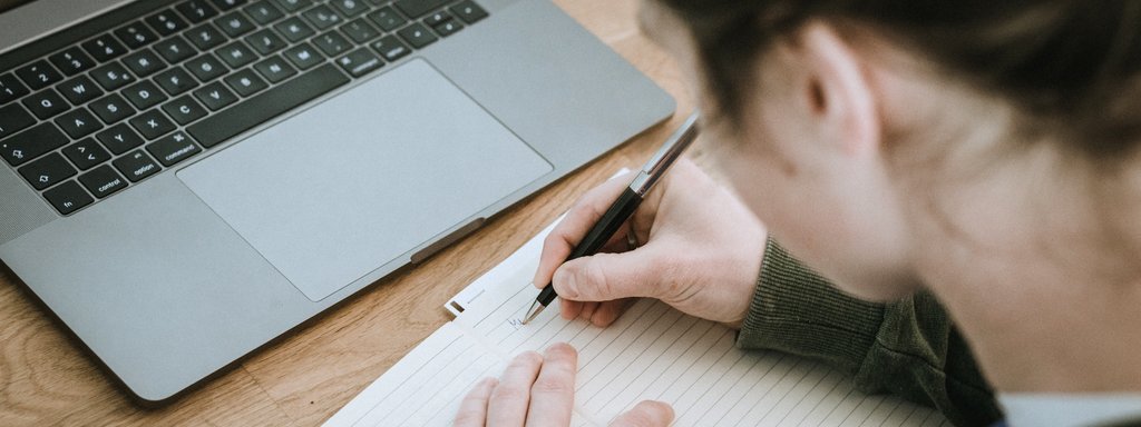Foto av ein student som sit ved ei datamaskin og skriv i ei notatblokk på bordet framfor seg. Vi ser personen over skuldra.