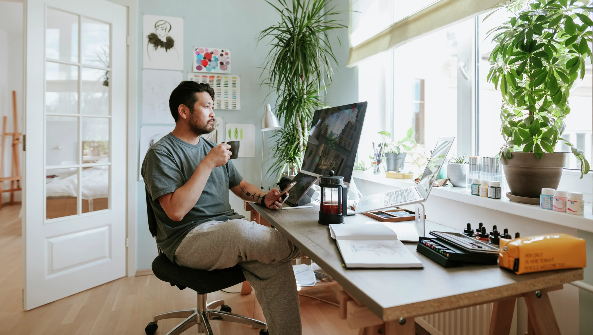 Gutt sitter ved en kontorpult hjemme med PC, mobil og en kaffekopp og ser ut av vinduet