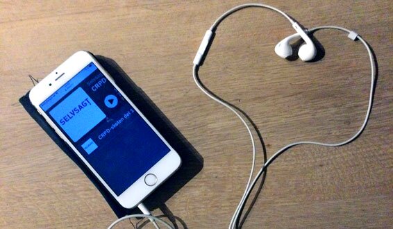 Mobiltelefon som viser Selvsagtpodden i displayet, ørepropper med ledninger som former et hjerte.