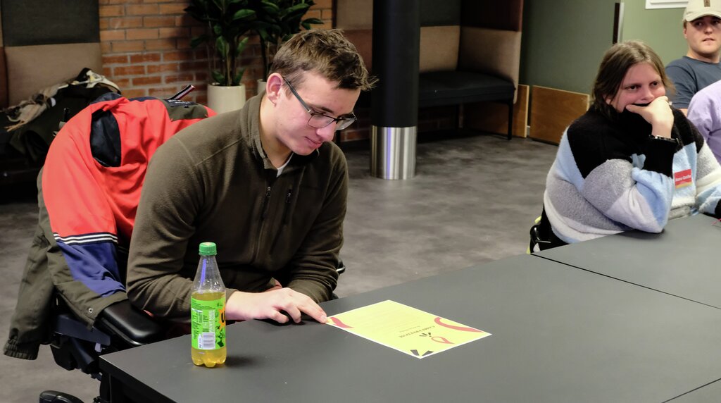 Andreas Melbye ser ned på sitt diplom som ligg framfor han på bordet. Han har briller, gråbrun genser og sit i elektrisk rullestol. 