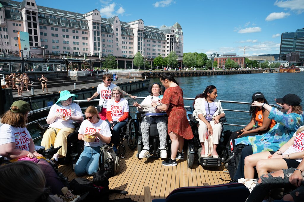 En smilende gjeng med og uten rullestoler på båten Arnøy i strålende solskinn. Flere har på seg t-skjorter der det står "Stolt, sterk, synlig!".