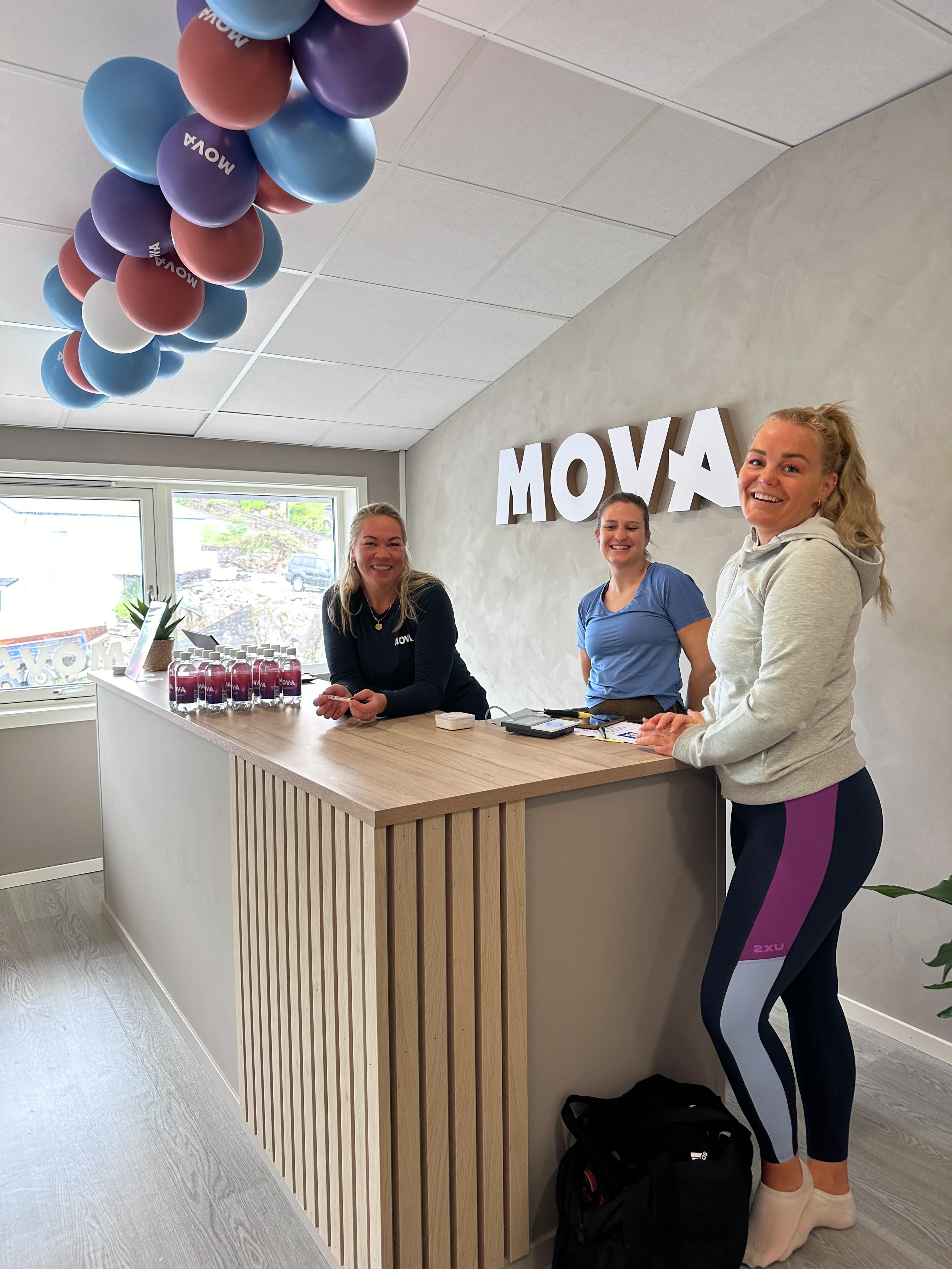 Ansatte og et medlem smiler ved resepsjonsdisken med MOVA-logo og ballonger på MOVA Mathopen.