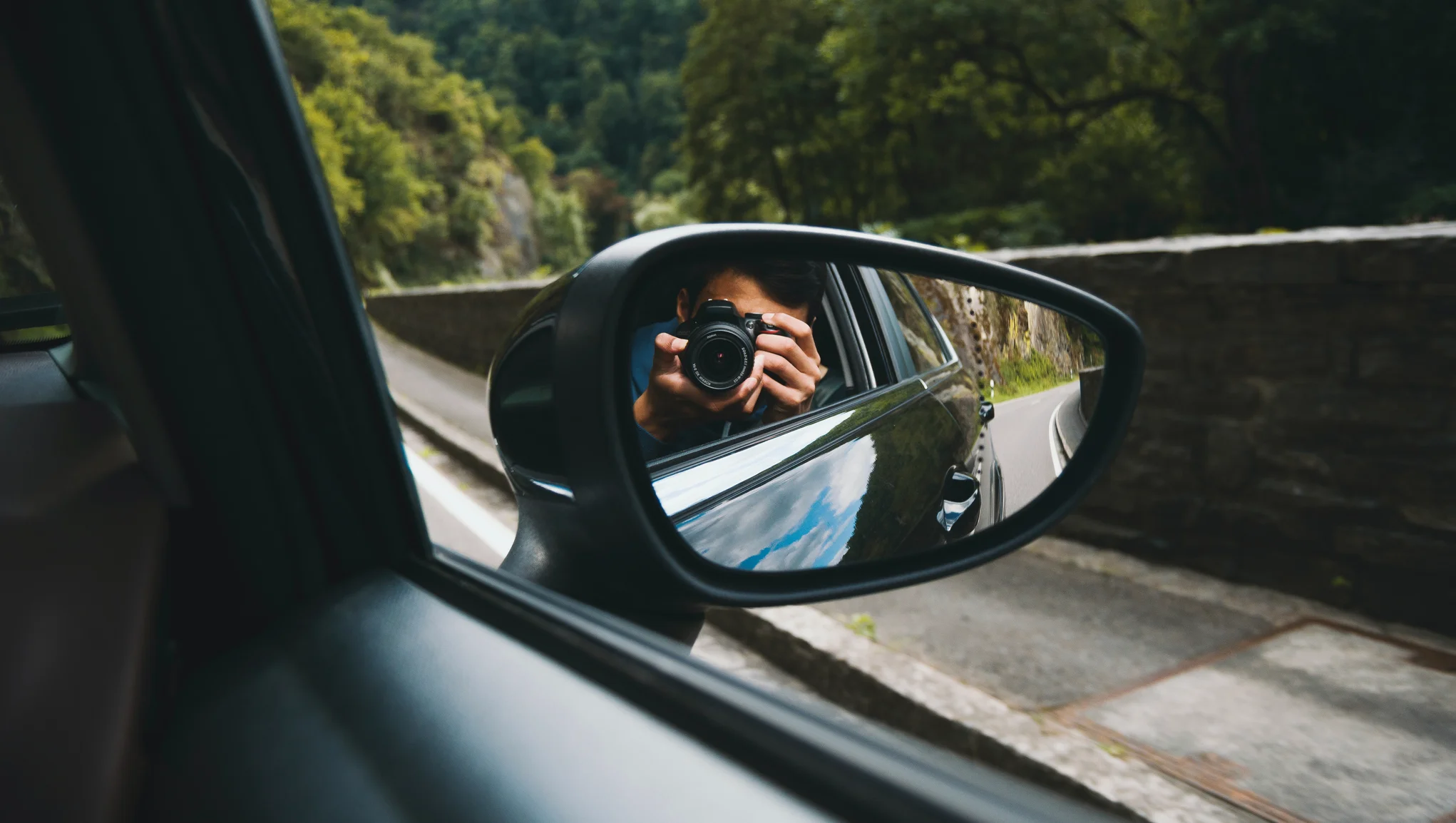 Elektromobilio keleivio vietoje sėdintis žmogus fotografuoja vaizdą už lango ir veidrodėlyje matosi jo kameros atspindys.