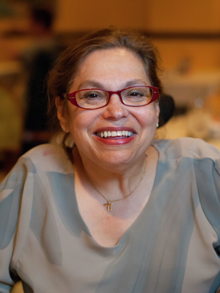 Judith "Judy" Heumann had a smile and a laugh that spread across her face. In the picture, she had her brown hair affixed with a tie, red glasses, light gray blouse, gold jewelery and red lipstick.