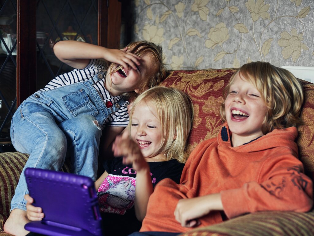 Tre leende barn i en sofa, ett holder et lilla nettbrett. De ser glade og underholdte ut.