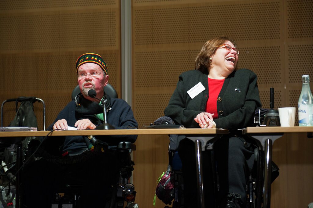 Kalle Könkkölä and Judy Heuman behind a panel table at a conference. Könkkölä has a hat with  patterns on it and glasses, Heumann has a red top, black jacket and is smiling at something outside the edge of the picture.
