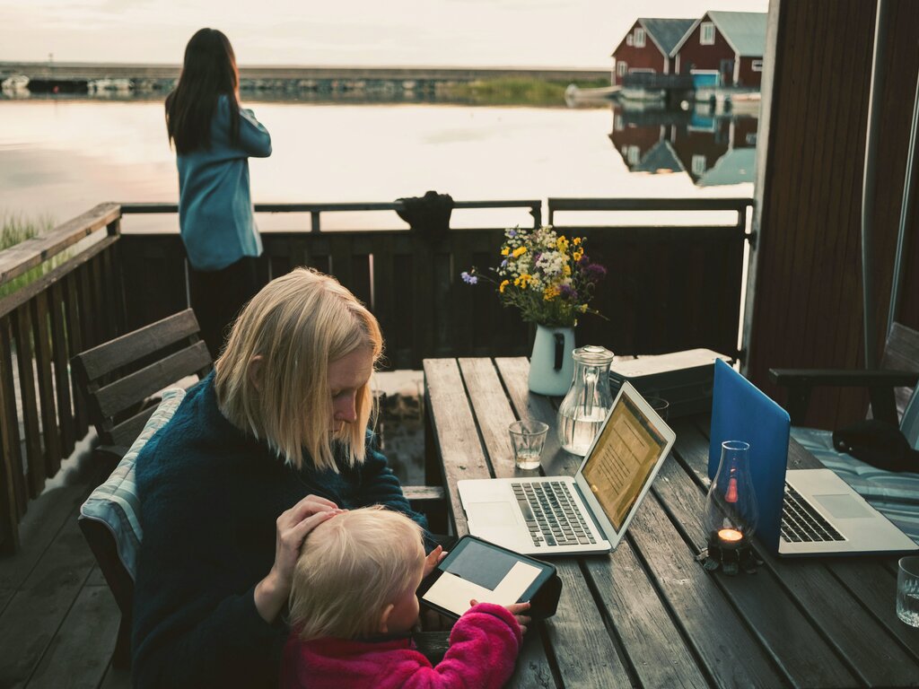 Kvinne som jobber på bærbar PC med barn på balkong med utsikt over vannet. En annen person stirrer på utsikten. Rustikk setting med blomster.