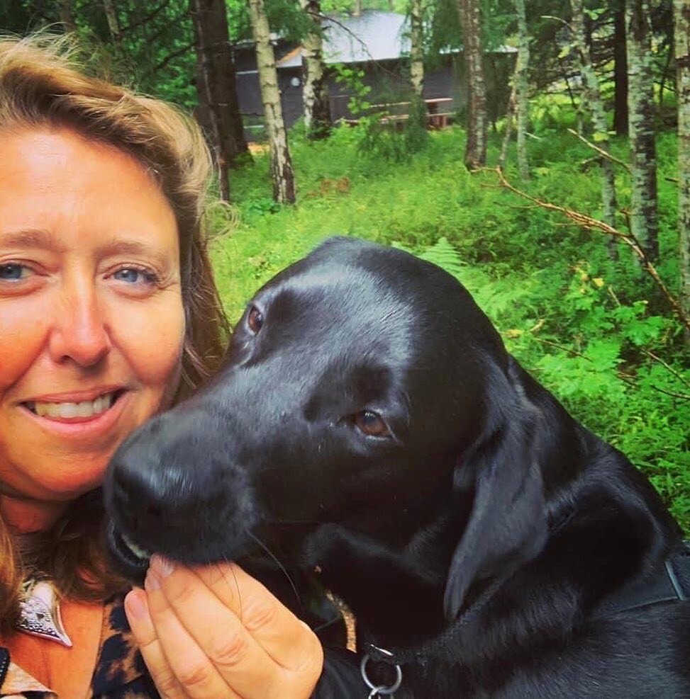 Heidi Anita Rahm og servicehunden Ivo. Rahm har lysebrunt hår, blå øyne, og hun smiler mens den svarte labradoren hennes tar et jafs av en godbit hun har i hånden.