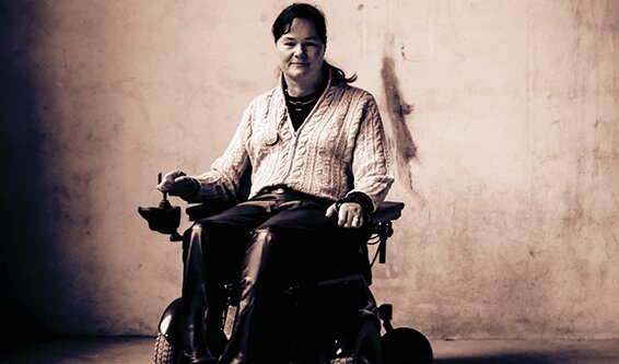 Et sepiafarget bilde av Vibeke Marøy Melstrøm i sin elektriske rullestol. Hun har strikkejakke på seg, et nokså nøytralt ansiktsuttrykk med unntak av et lite smil, og sitter foran en bar murvegg.
