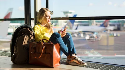 Nainen selaa puhelinta ja istuu lentokentän lattialla ikkunan vieressä