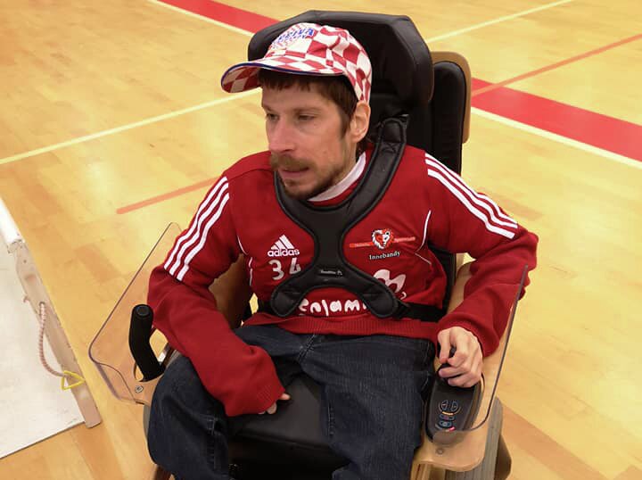 Karl Wilhelm Faye-Hansen i rød lagdrakt på el-innebandy. Han har en rød og hvitrutete caps på hodet og brunt hår og skjegg. Her sitter han også i rullestolen han bruker spesifikt til idretten. 