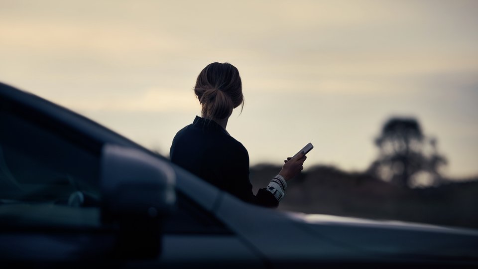 Kvinna i motljus står med mobiltelefon i handen framför en Volvo