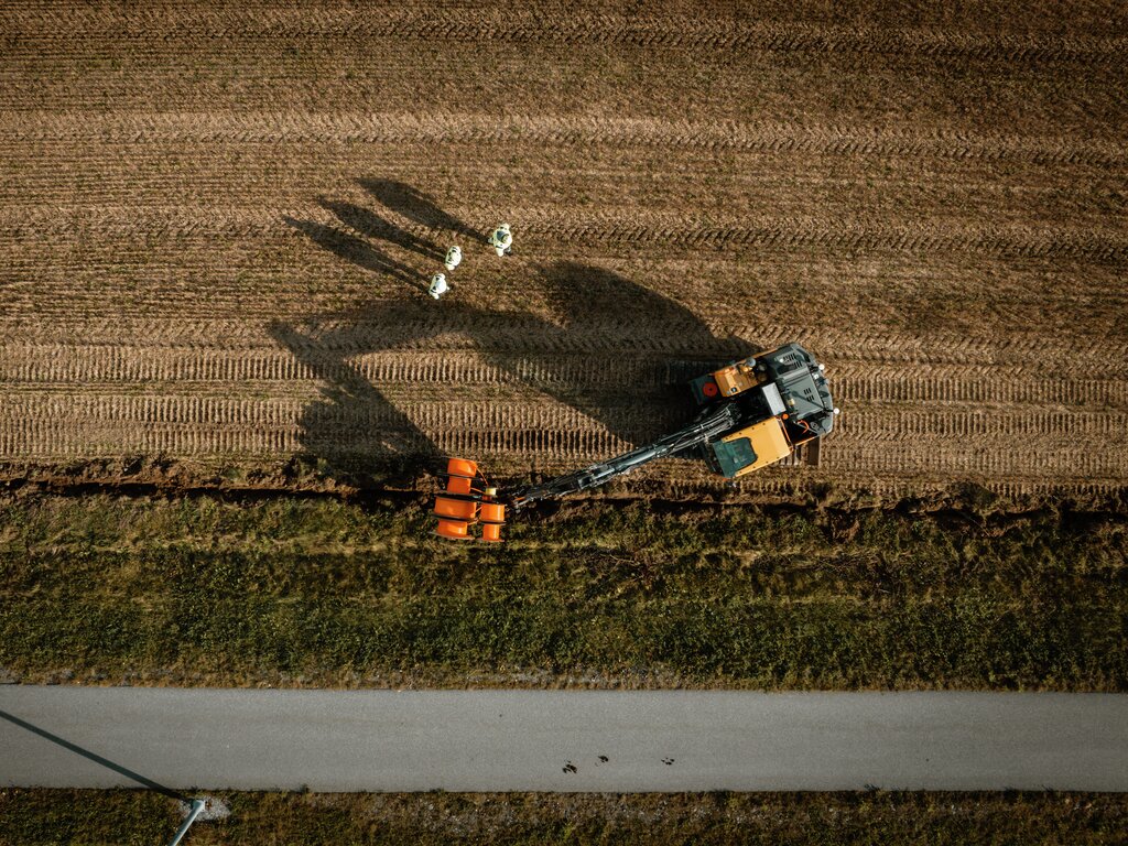 Luftfoto av traktor som presser høy på jordet og kaster lange skygger. Baller og betongsti er synlige i nærheten.