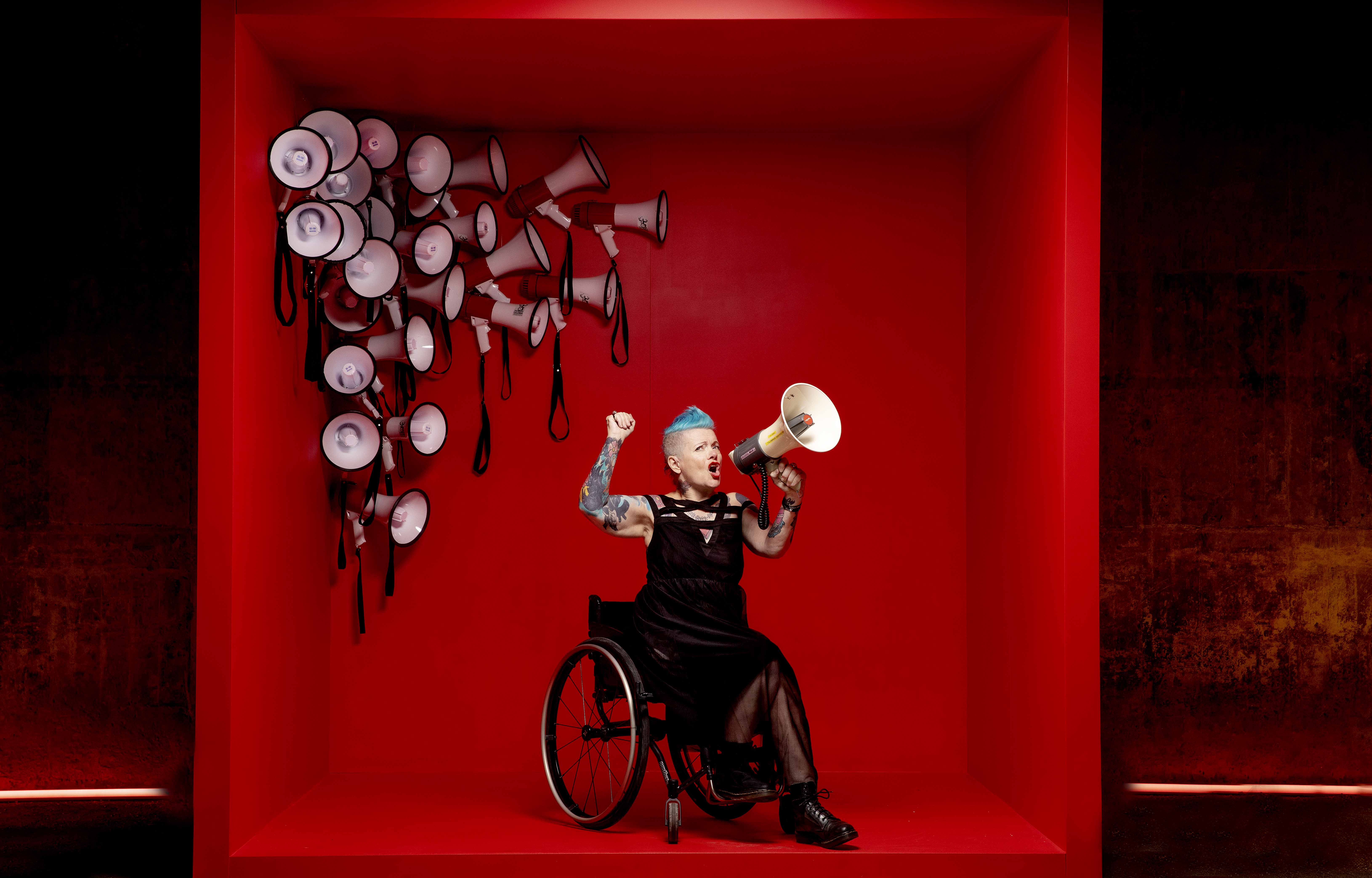 (1) Helle-Viv i en stor rød boks, med hvite og røde roperter i venstre hjørne. Helle-Viv sitter i en rullestol, med svarte klær, og roper inn i en ropert. Hun har mange tatoveringer, og blått kult hår. 