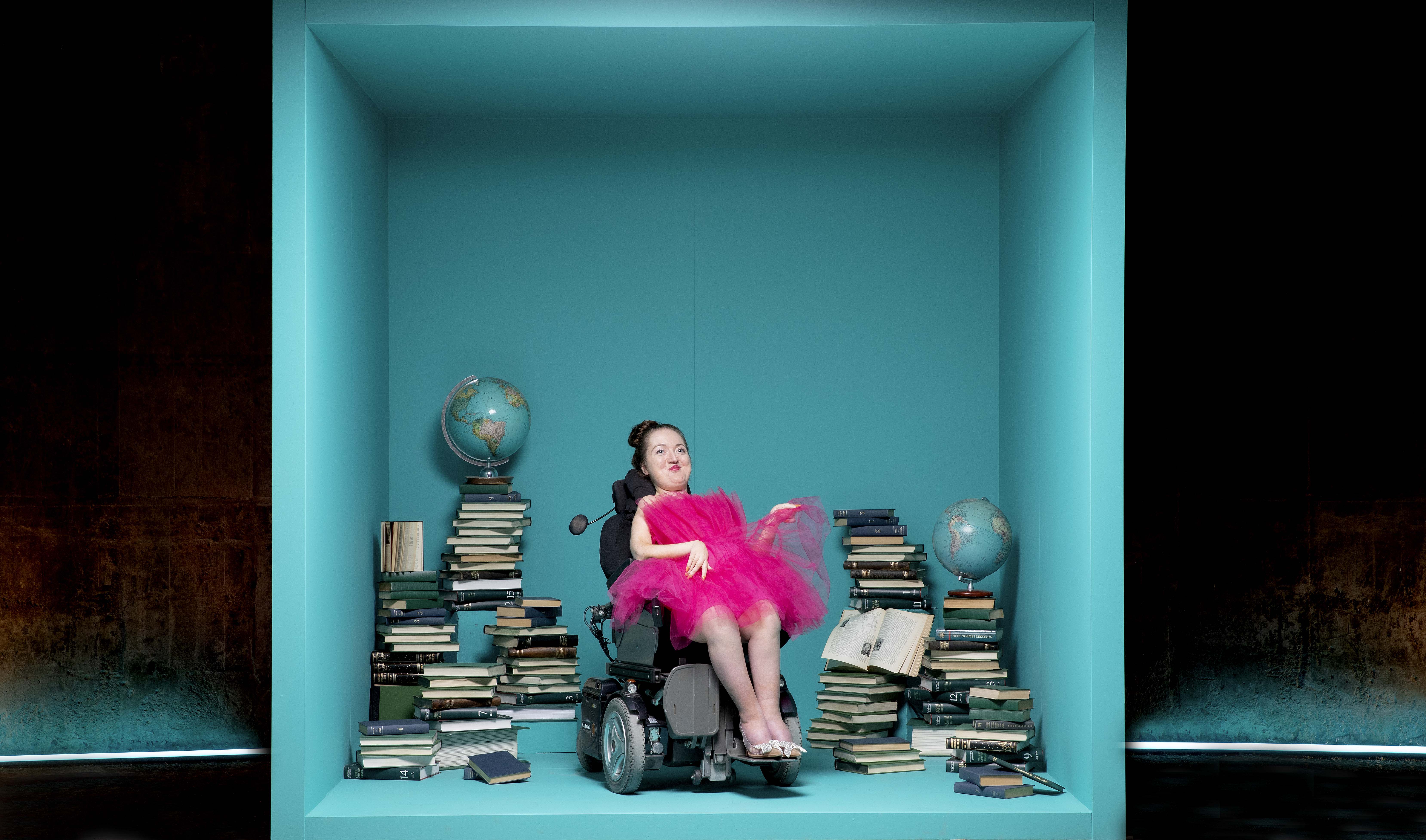 (1) Ingeborg sitter i sin rullestol inne i en stor blå boks. Hun har på seg en rosa tyllkjole. I boksen er det globus og mange bøker. 