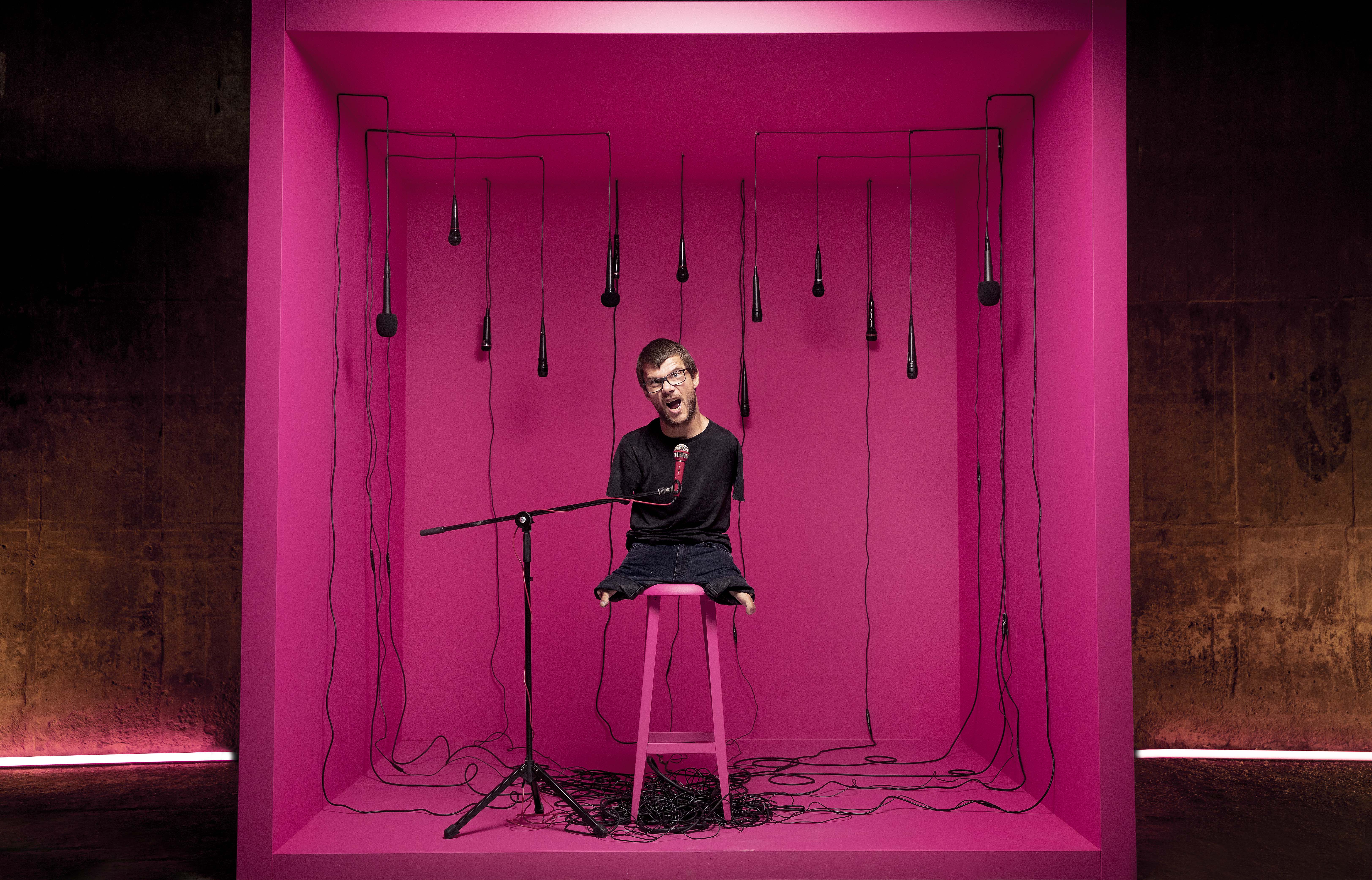 (1) Sebastian sitter inne i en rosa stor boks, med mikrofon han snakker inn i. Han sitter på en tamburrett og det er mikrofoner hengende ned fra taket. 