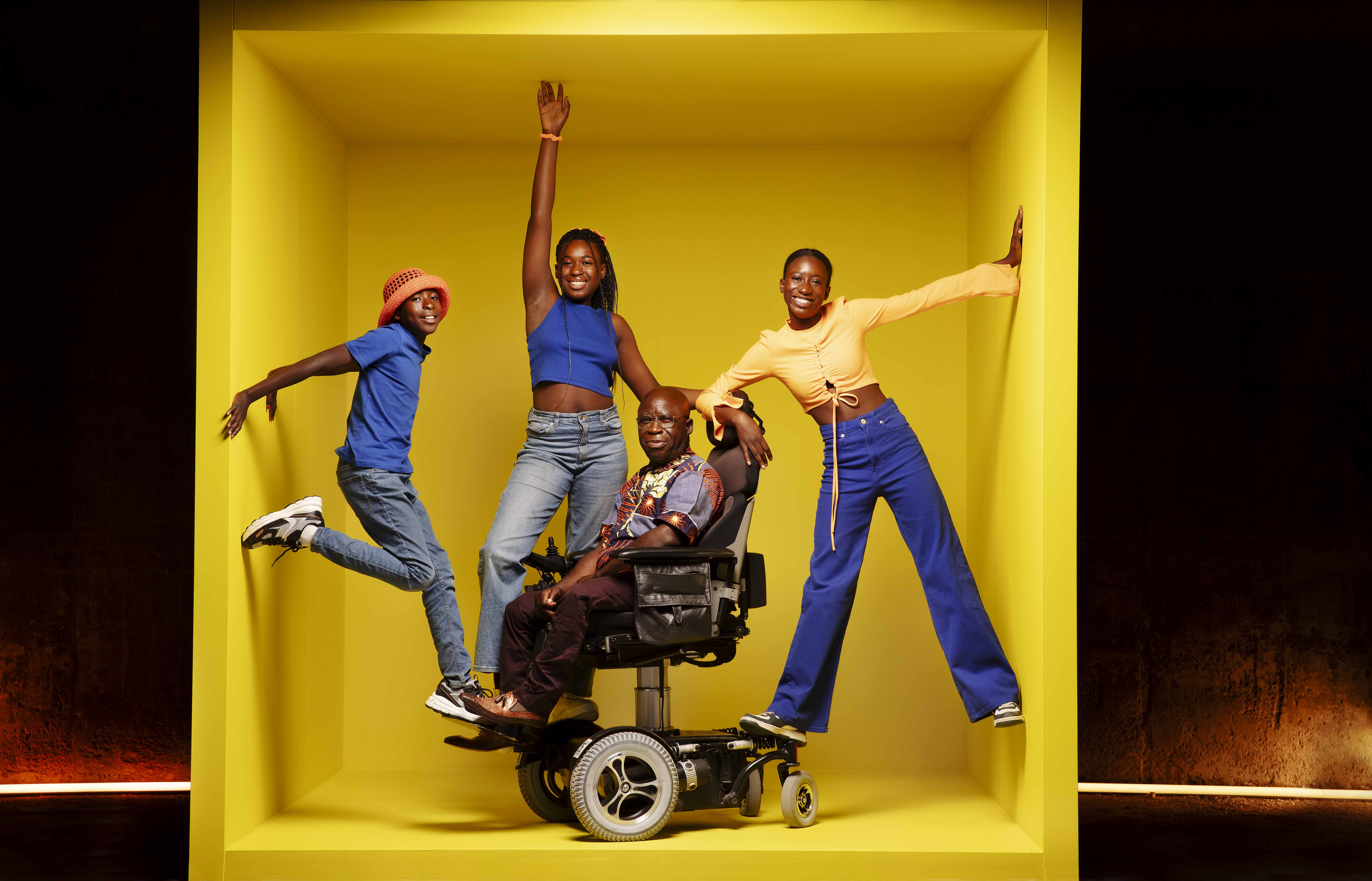 (1) Bihayo og tre av barna sine inne i en stor gul boks. Bihayo sitter i elektrisk rullestol, klærne til menneskene er fargerike i blå,brun og gul-nyanser. Barna balanserer på rullestolen. 