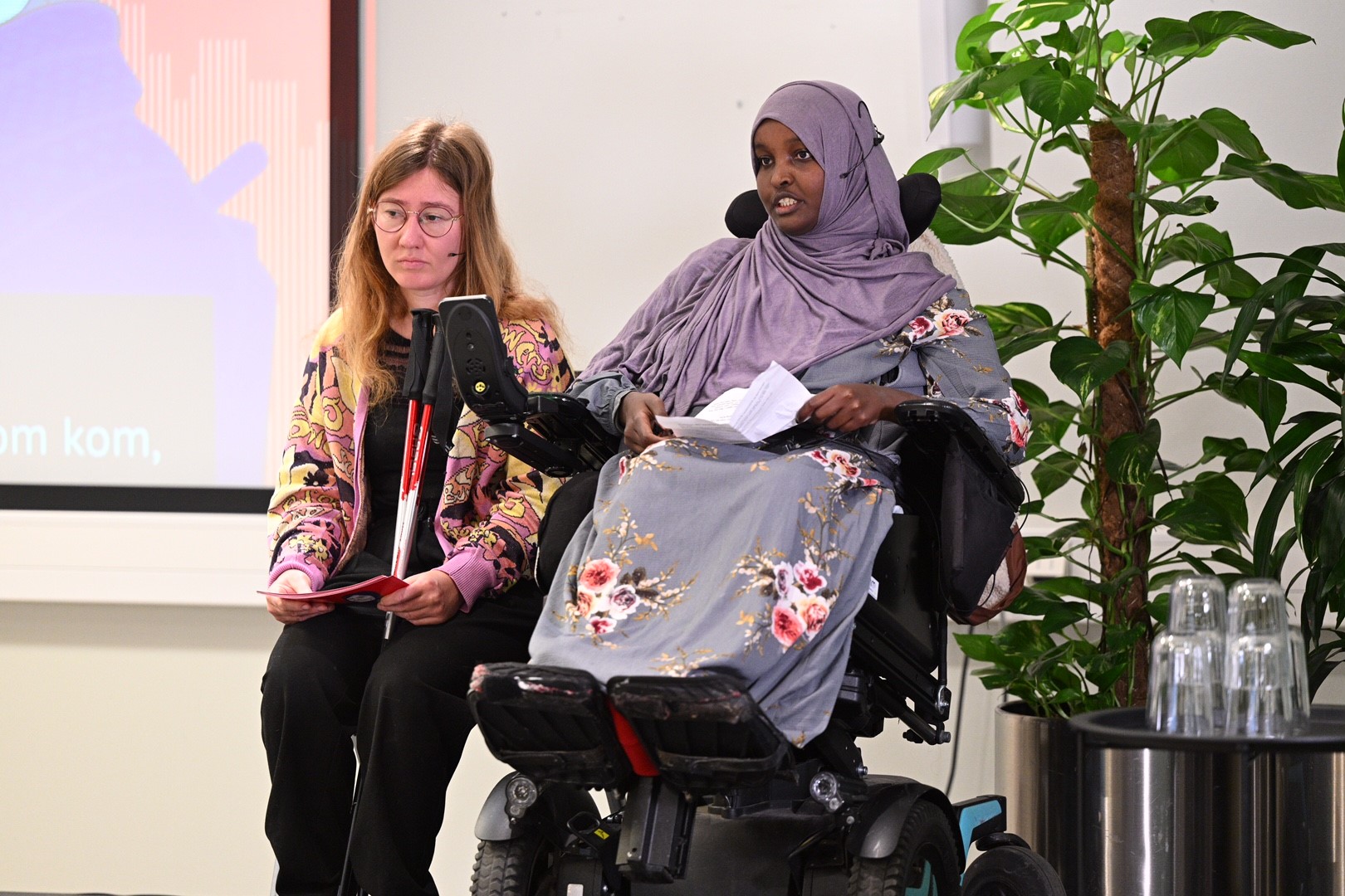 Habibo Mahammad Mahmud sitter på scenen i en elektrisk rullestol sammen med Martine Eliasson. Habibo har på seg en lilla hijab og blåblomstret kjole. Hun har mørk hud og brune øyne, og leser fra et ark.