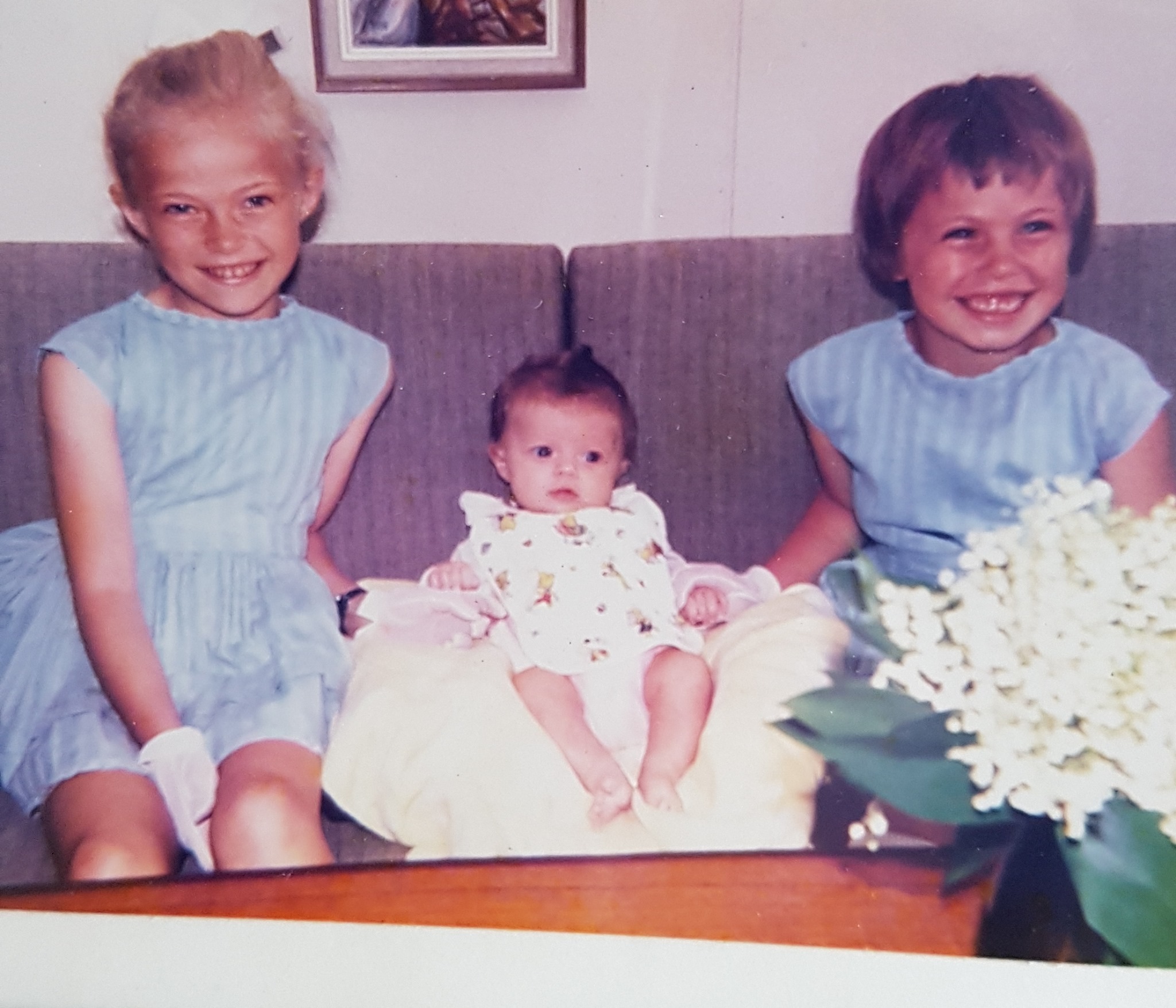 Helle-Viv og storesøstrene som barn. Helle-Viv sitter i midten og er minst, iført en hvit kjole. De to storesøstrene sitter på hver sin side like blå kjoler. Alle smiler til kameraet. 