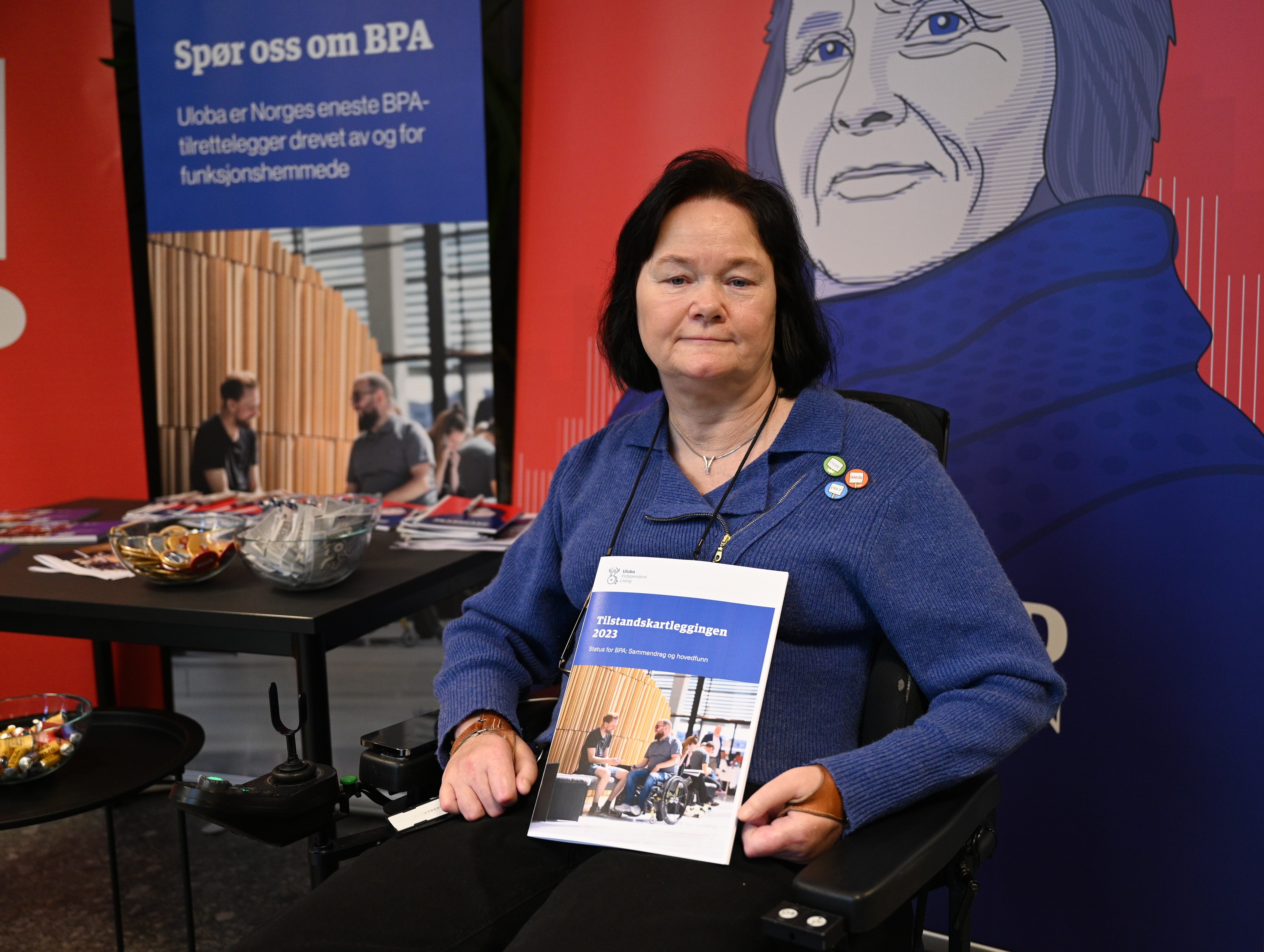 Vibeke Marøy Melstrøm foran en Skansgård-parole. Hun holder opp Tilstandskartleggingen 2023. Hun har mørkt skulderlangt hår, blå overdel, svart bukse og  blå øyne. Hun sitter i en elektrisk rullestol.