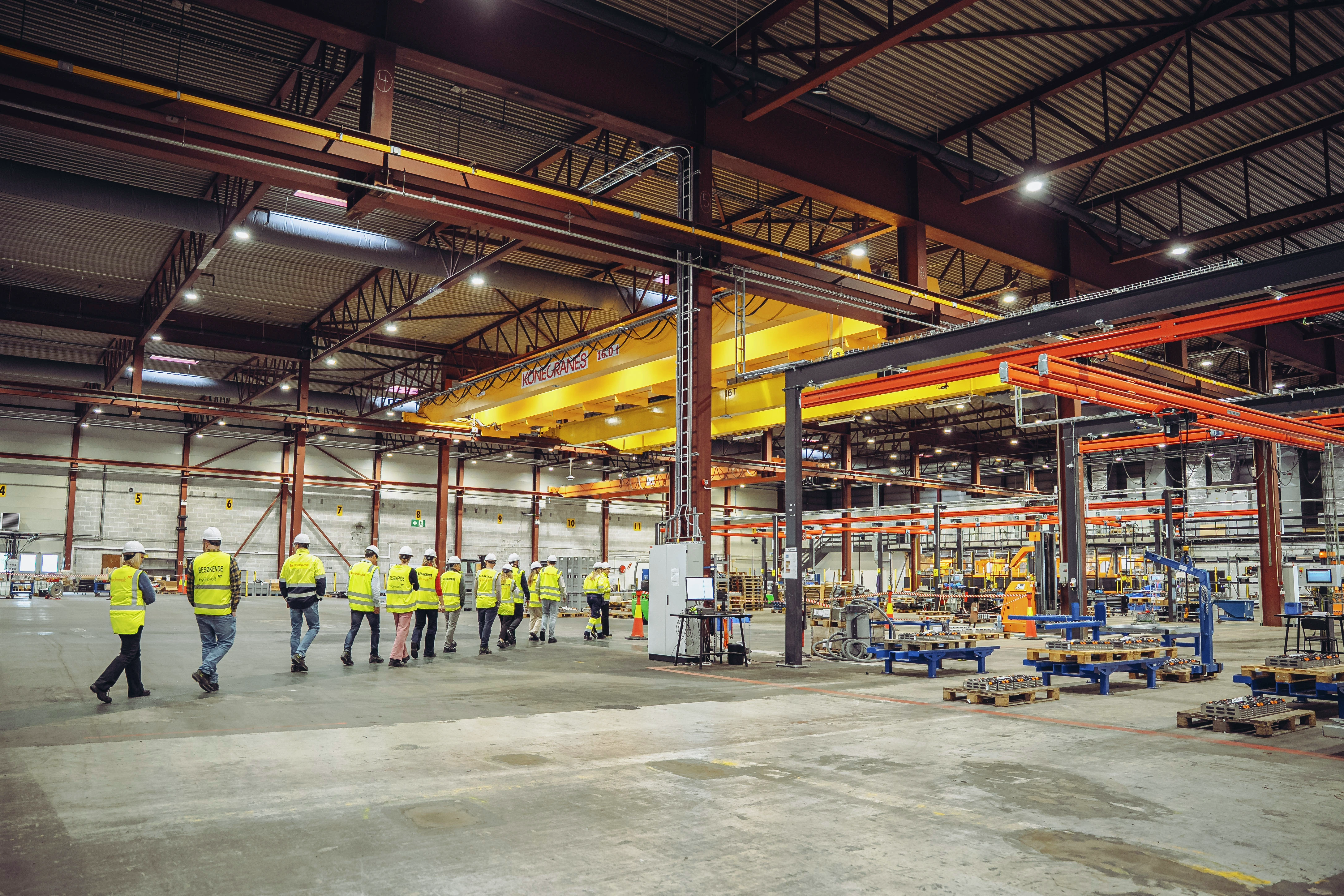 Interiør av industrilager med arbeidere i gule sikkerhetsvester, traverskraner, maskiner og utstyr på et betonggulv.