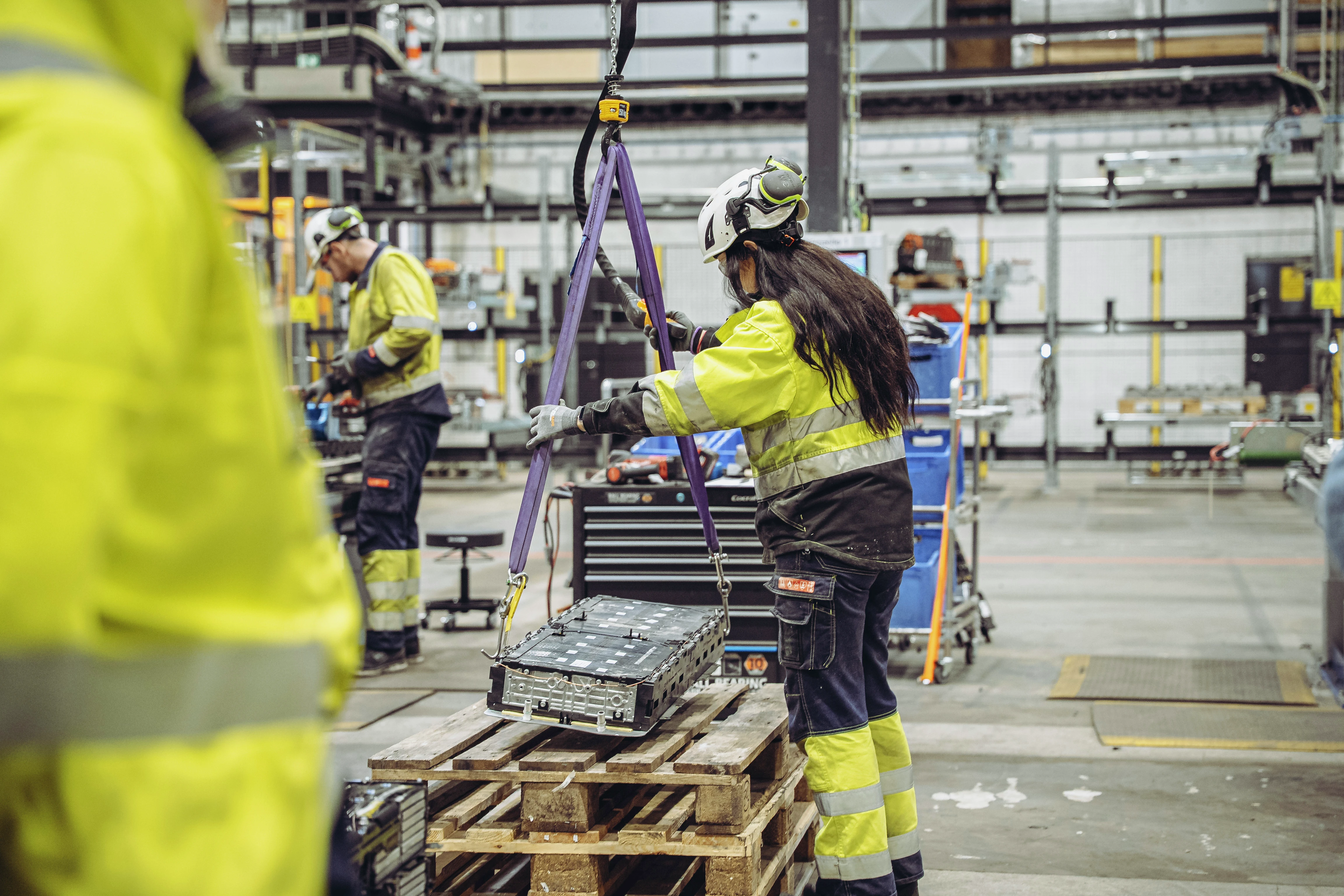 Arbeidere i sikkerhetsutstyr betjener maskiner i industrianlegg. Kvinne justerer løftestropper på pall med metallkomponent.