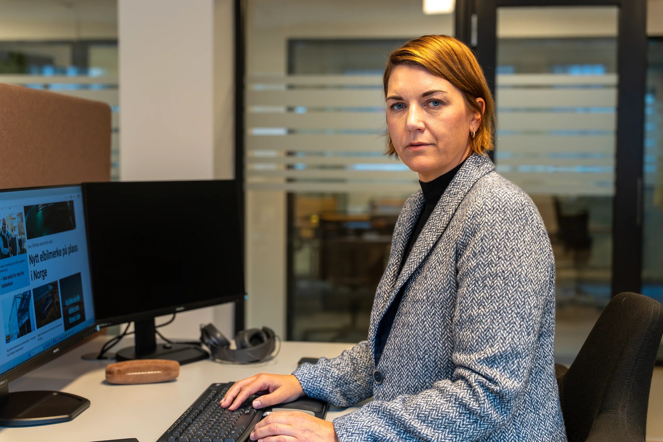 En profesjonell kvinne i en grå blazer sitter ved et skrivebord med to skjermer på et moderne kontor.