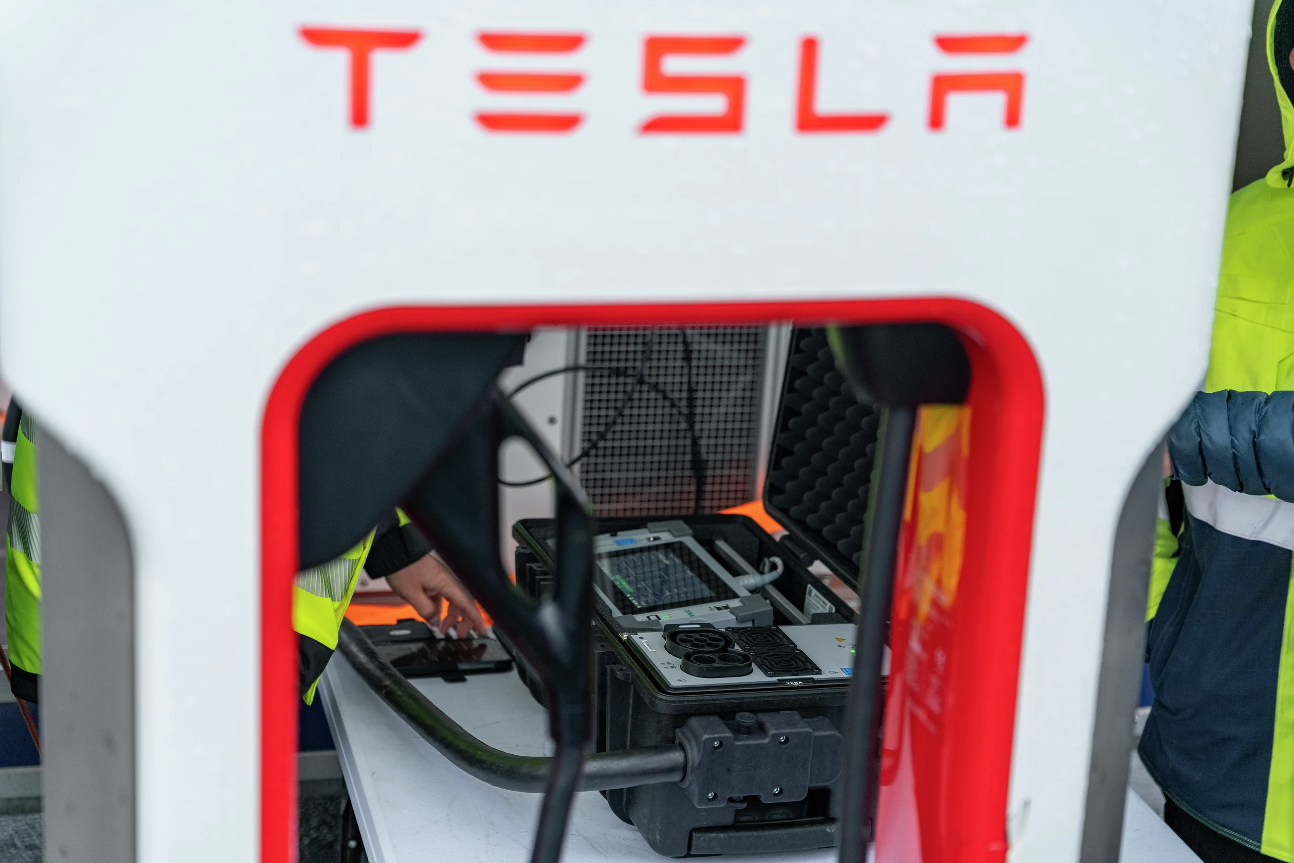 Tesla Supercharger-stasjonen er åpen for vedlikehold, med teknikere i refleksvester som jobber med interne elektriske komponenter.