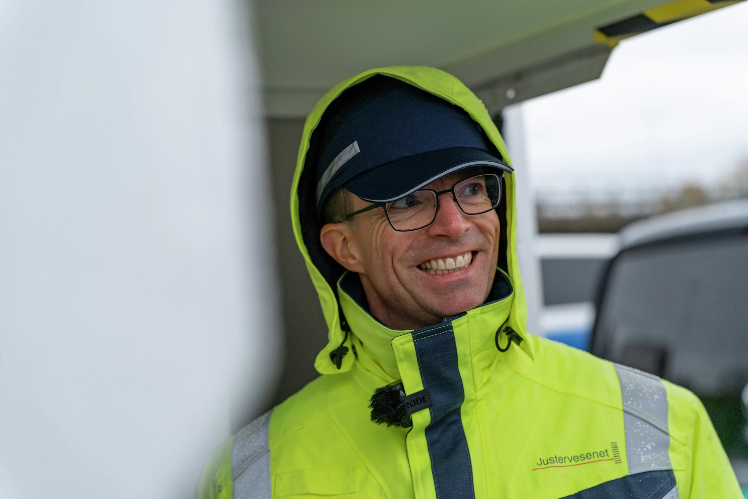 En smilende mann med briller og en gul refleksjakke med Justervesenet-logo og hette oppe.