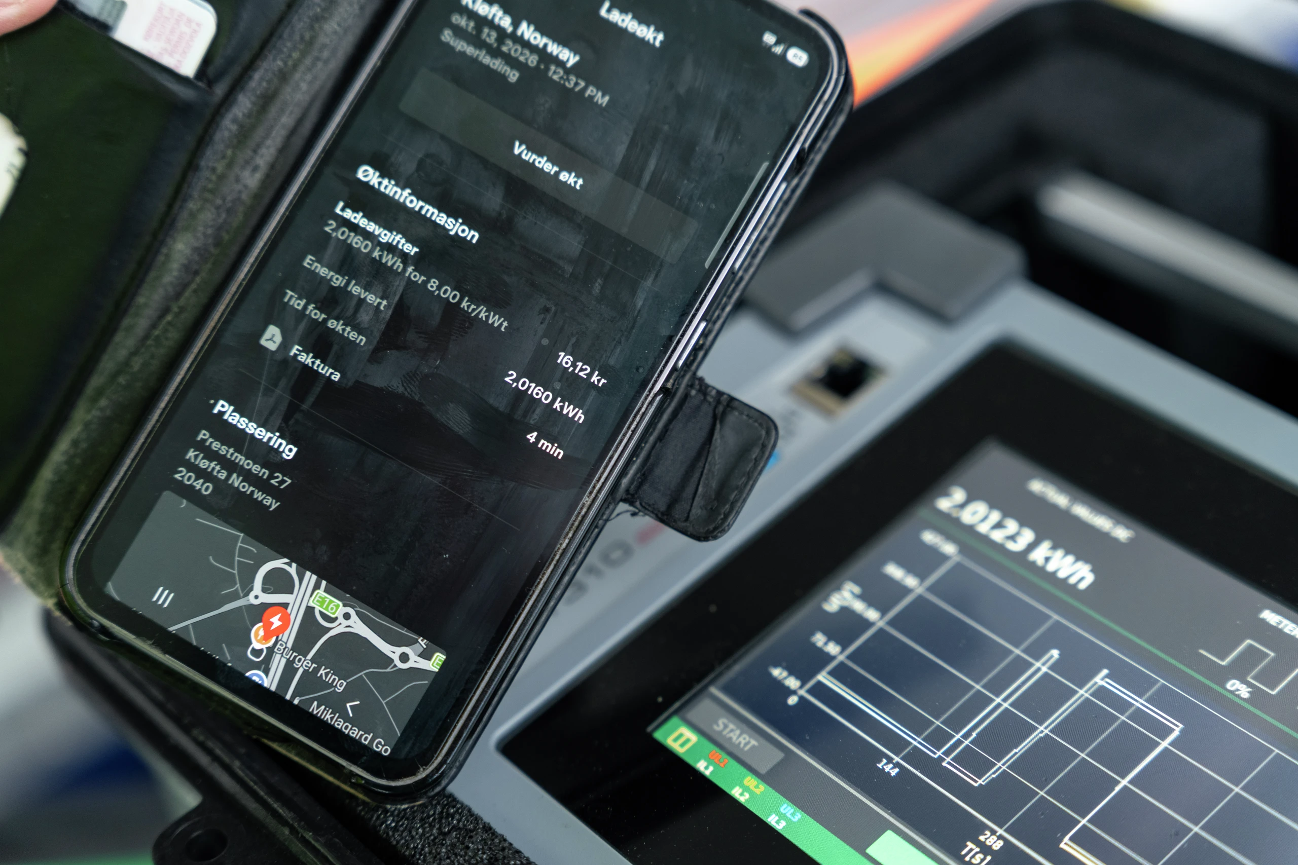 En smarttelefon montert i en bil som viser en ladeapp for elbiler med norsk tekst og detaljer om ladestasjonen.