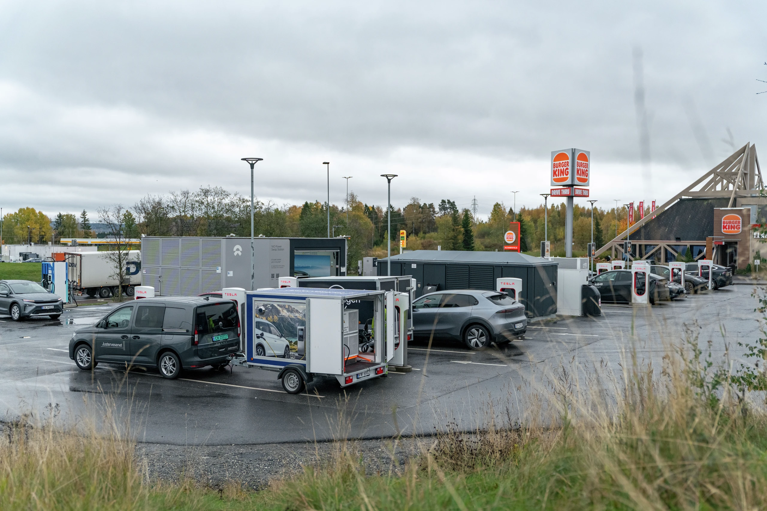 En travel Burger King-parkeringsplass med Tesla Superchargers, NIO-ladestasjoner og diverse elbiler på en overskyet dag.