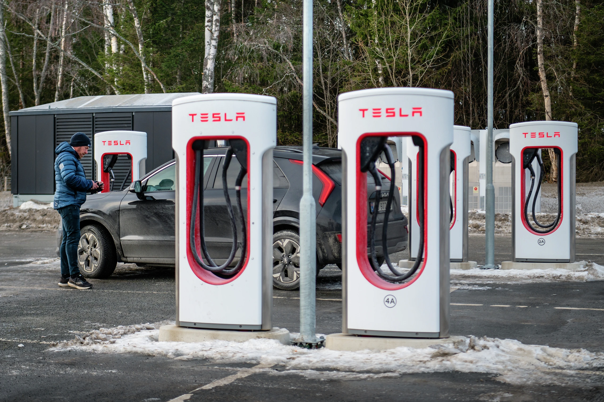 Tesla Supercharger-stasjon med flere ladeenheter. Person i blå jakke lader mørk SUV. Snødekt terreng, trær i bakgrunnen.