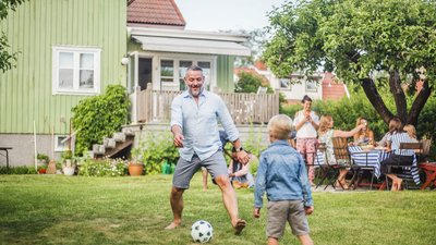 Mies ja lapsi pelaavat jalkapalloa omakotitalon nurmikolla.
