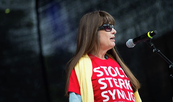 Berit Vegheim foran en mikrofon og mørk bakgrunn. Hun har på seg solbriller, gult skjerf og rød T-skjorte med Ulobas slagord "Stolt, sterk, synlig" på.