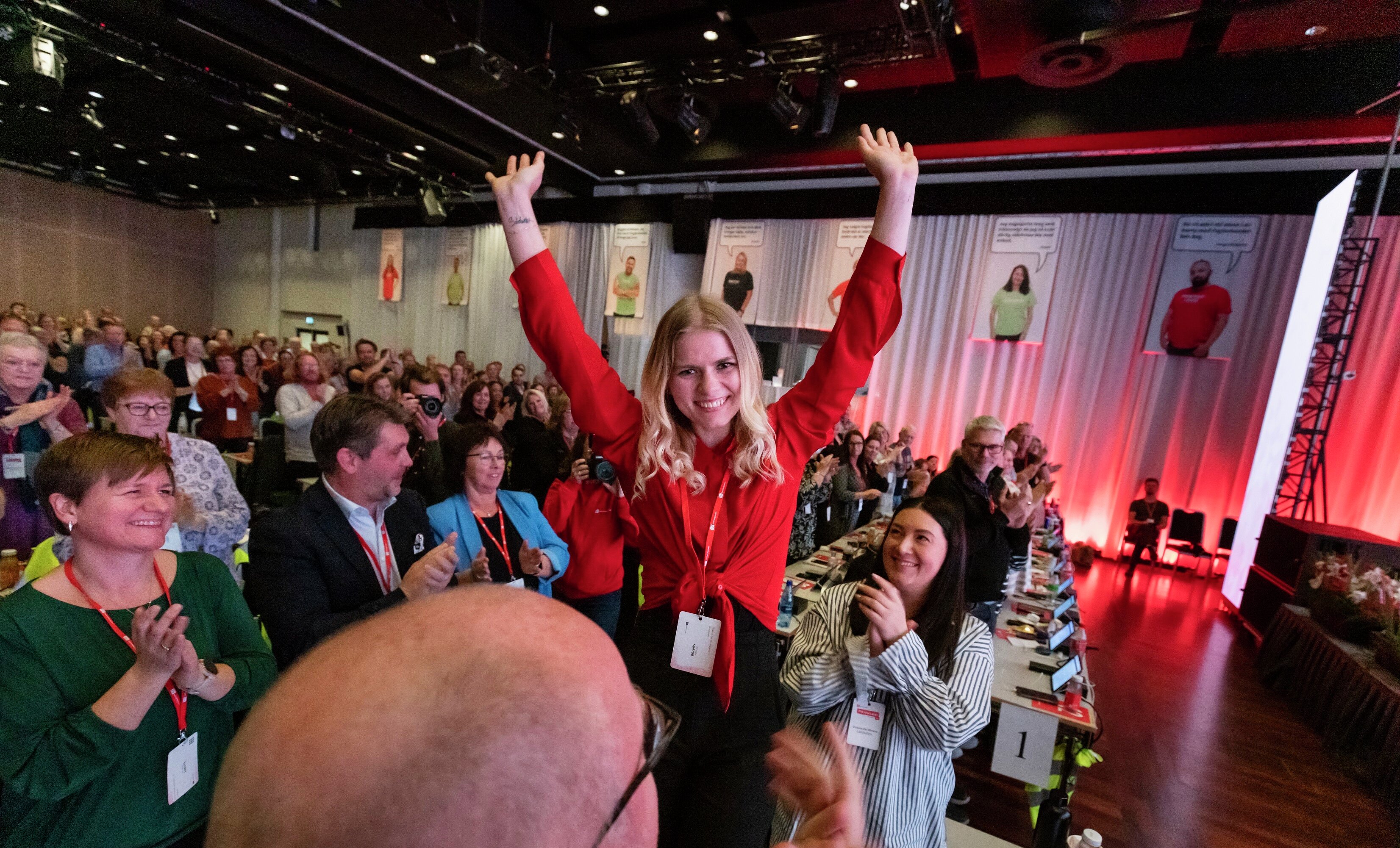 Ung kvinne med rød skjorte, smiler og står med armene i været for å feire seieren. Rundt henne står hundrevis av folk på Fagforbundets landsmøte og klapper.