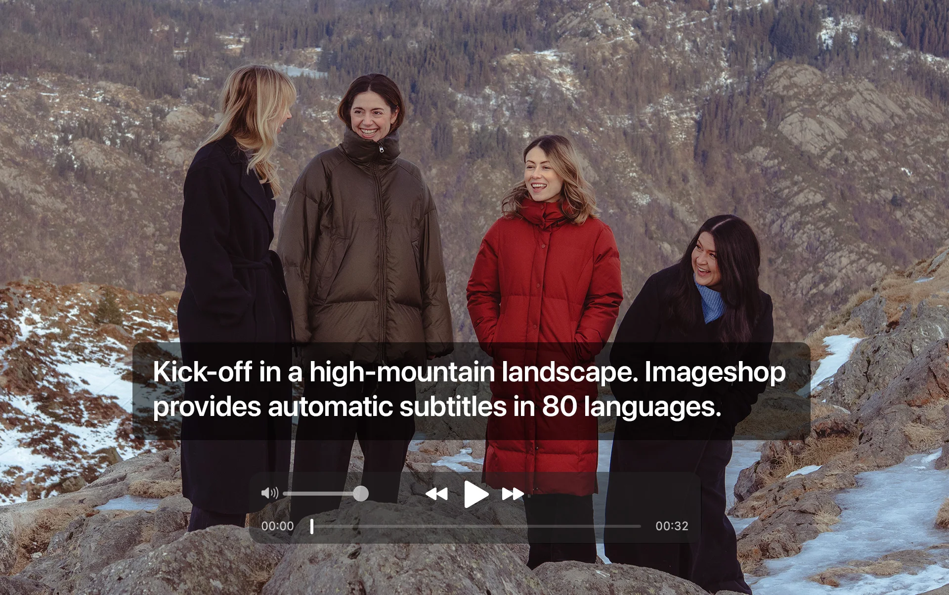Four women in winter coats laugh together in a rocky mountain landscape with patches of snow.