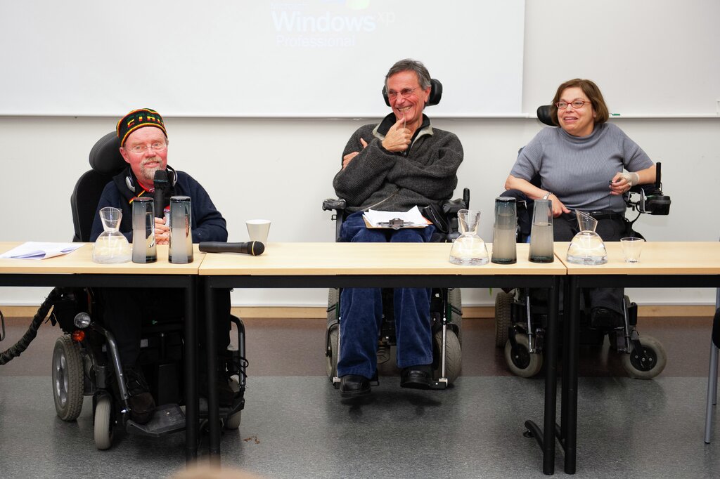 Kalle Könkkölä, Adolf Ratzka og Judy Heumann i eit lattermildt augeblikk på konferanse. Ratzka og Heumann smiler mot Könkkölä som snakkar inn i ein mikrofon. 