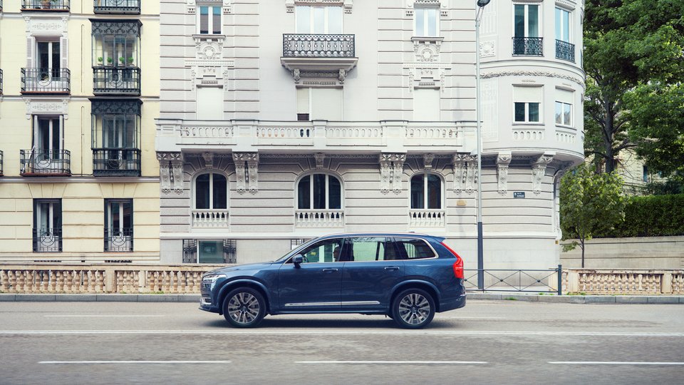 Blå Volvo XC90 åker utmed gata i stadsmiljö med stenhus i bakgrunden