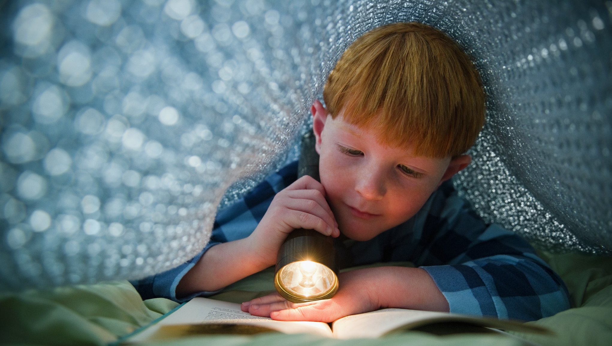 En pojke läser med ficklampa under ett täcke