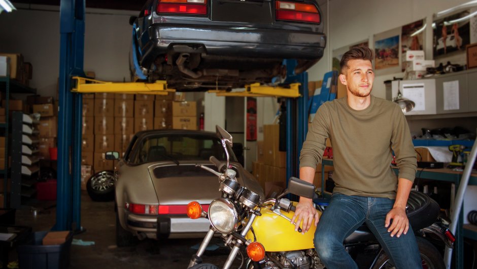 Vyras garaže atsirėmęs į geltoną motociklą, už jo nugaros du automobiliai.