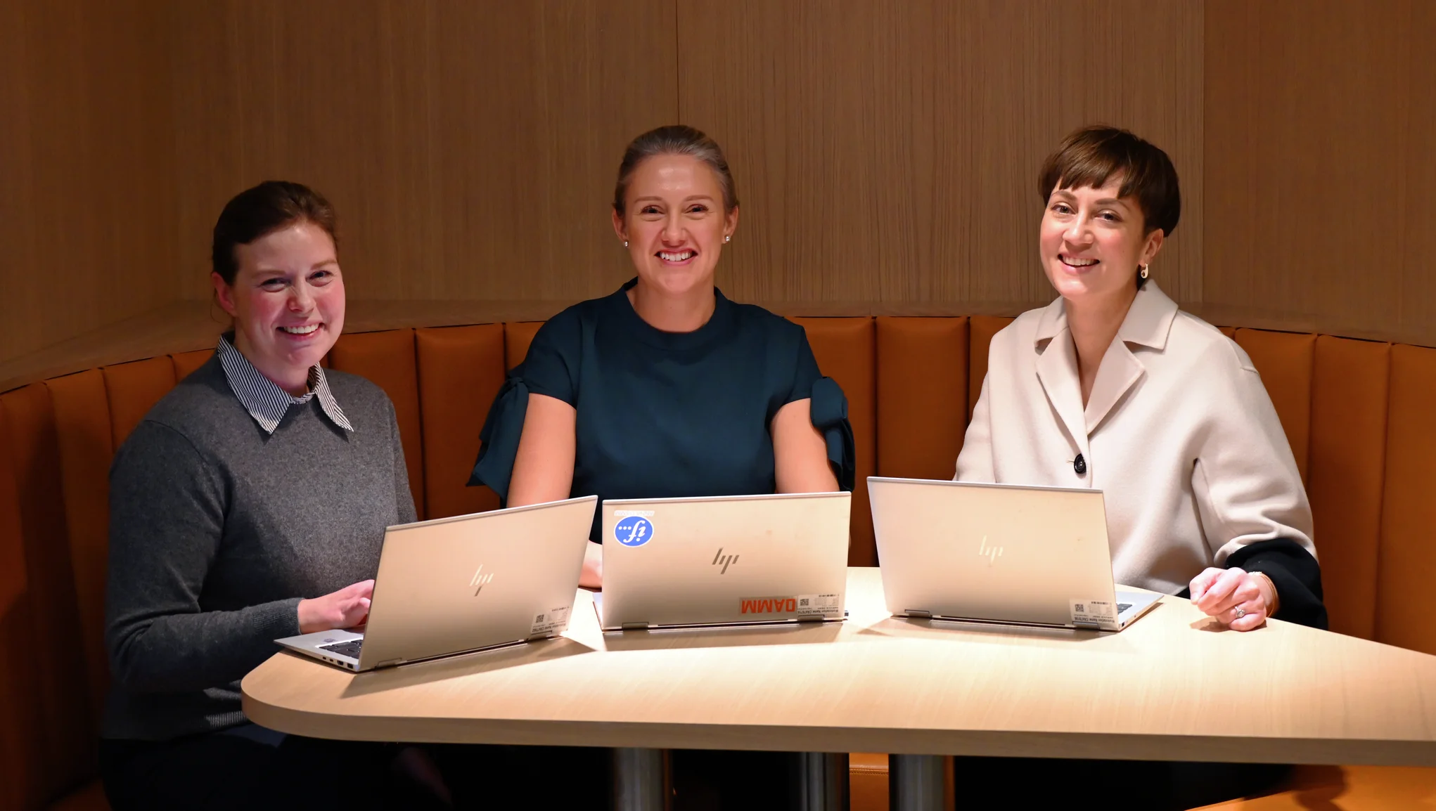 Kolme hymyilevää vastuullisuusviestijää vierekkäin pyöreän pöydän ääressä kannettavat tietokoneet edessään