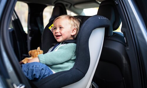Skrattande pojke med gosedjur i knät sitter i bakåtvänd bilbarnstol
