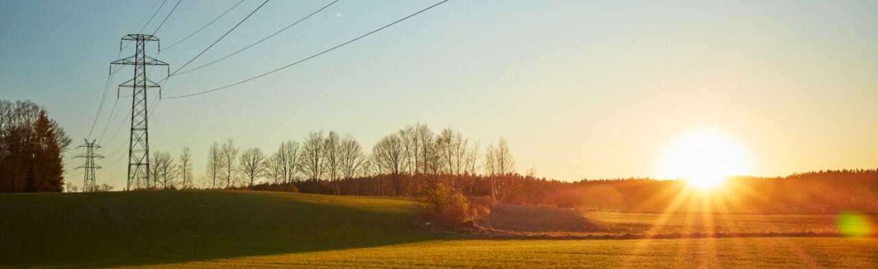 Solnedgang over landlig jorde med elektriske overføringstårn og kraftledninger som strekker seg over en klar himmel.