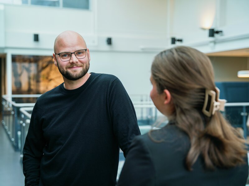 En mann ser smilende inn i kamera, en kvinne står med ryggen til
