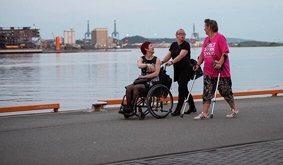Kvinne i rullestol trilles av assistent ved siden av kvinne med krykker og iført rosa Stolt Sterk Synlig-t-skjorte, langs kaia ved Bjørvika