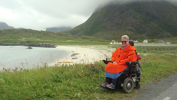 Bildet viser Sonja Tobiassen i knall oransje anorakk ute i et nordnorsk sommerlandskap. Hun sitter ved en hvit strand på Hovden i hjemstedet Vesterålen.